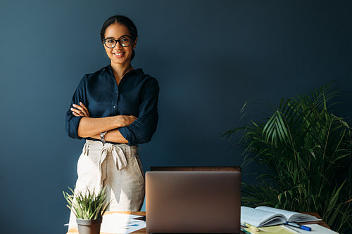 Smiling woman with arms crossed stands at her workplace in home office