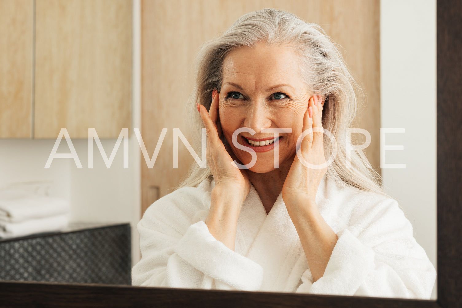 Smiling woman with grey hair massaging her face with her fingers. Aged female looking at her reflection in a mirror in the morning.