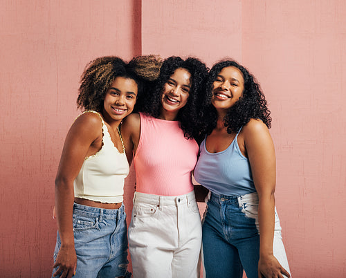 Three positive women with curly hair standing together against a pink wall and looking at the camera