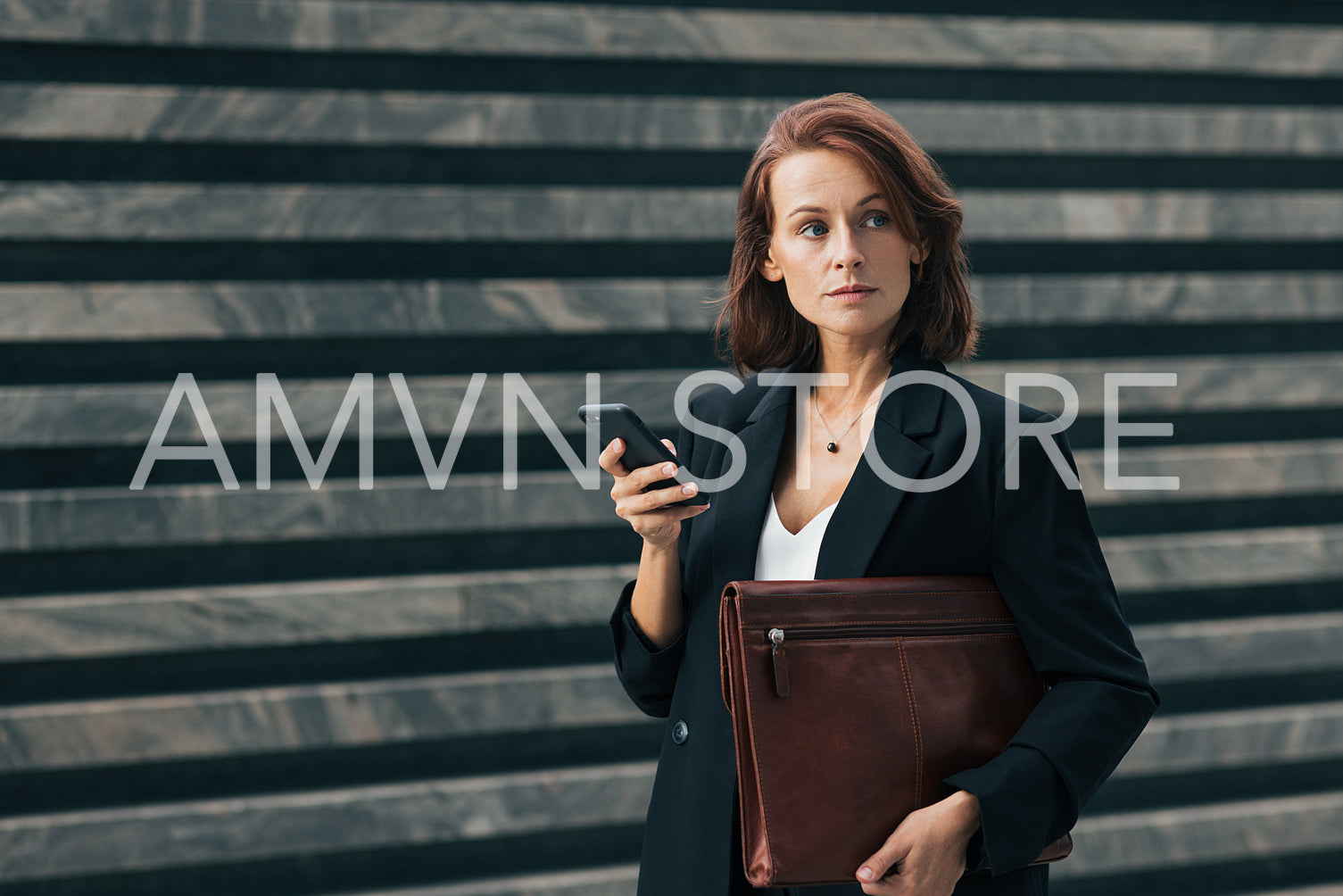 Confident businesswoman with ginger hair. Middle-aged woman holding smartphone and looking away. Female in formal clothes holding leather folder outdoors.