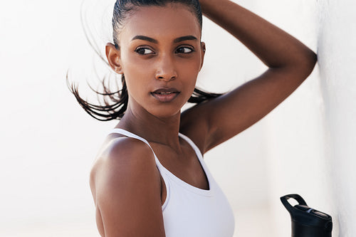 Portrait of a beautiful sportswoman relaxing at a white wall. Young female leaning wall looking away while taking a break.