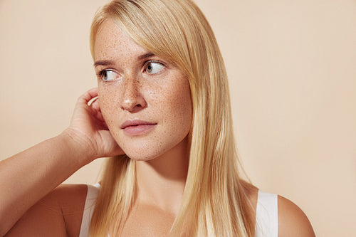 Close-up of freckled female looking away while adjusting her blond hair in studio. Portrait of a beautiful woman with perfect smooth skin.