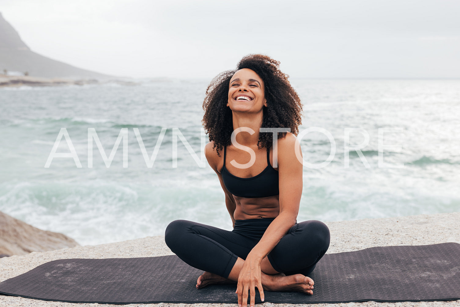 Laughing woman with curly hair sitting on mat by ocean. Female relaxing during yoga training outdoors.