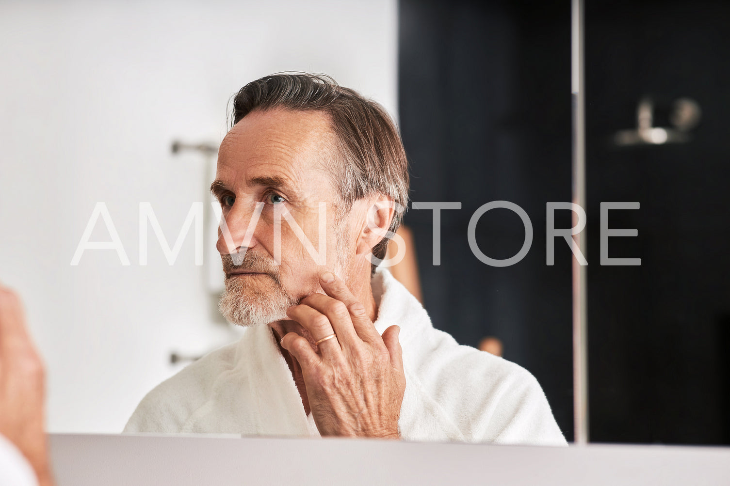 Senior man touching her face in bathroom in from of a mirror. Male in bathrobe looking at his reflection inspecting skin.	