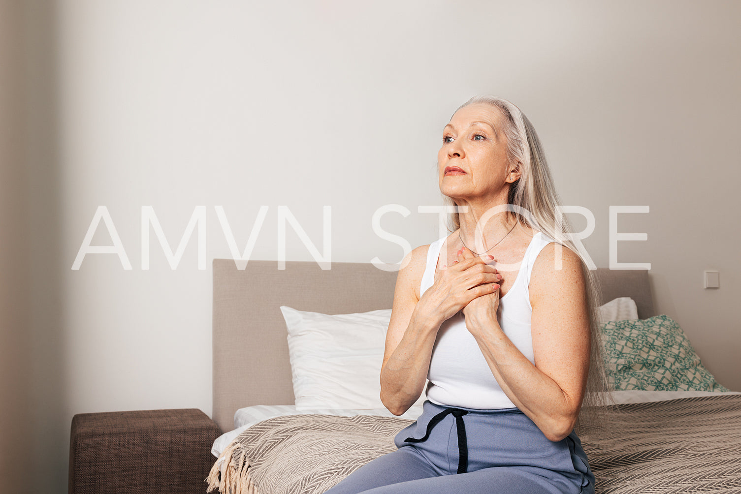 Worried senior woman sitting on a bed. Sad adult female with hands on chest.