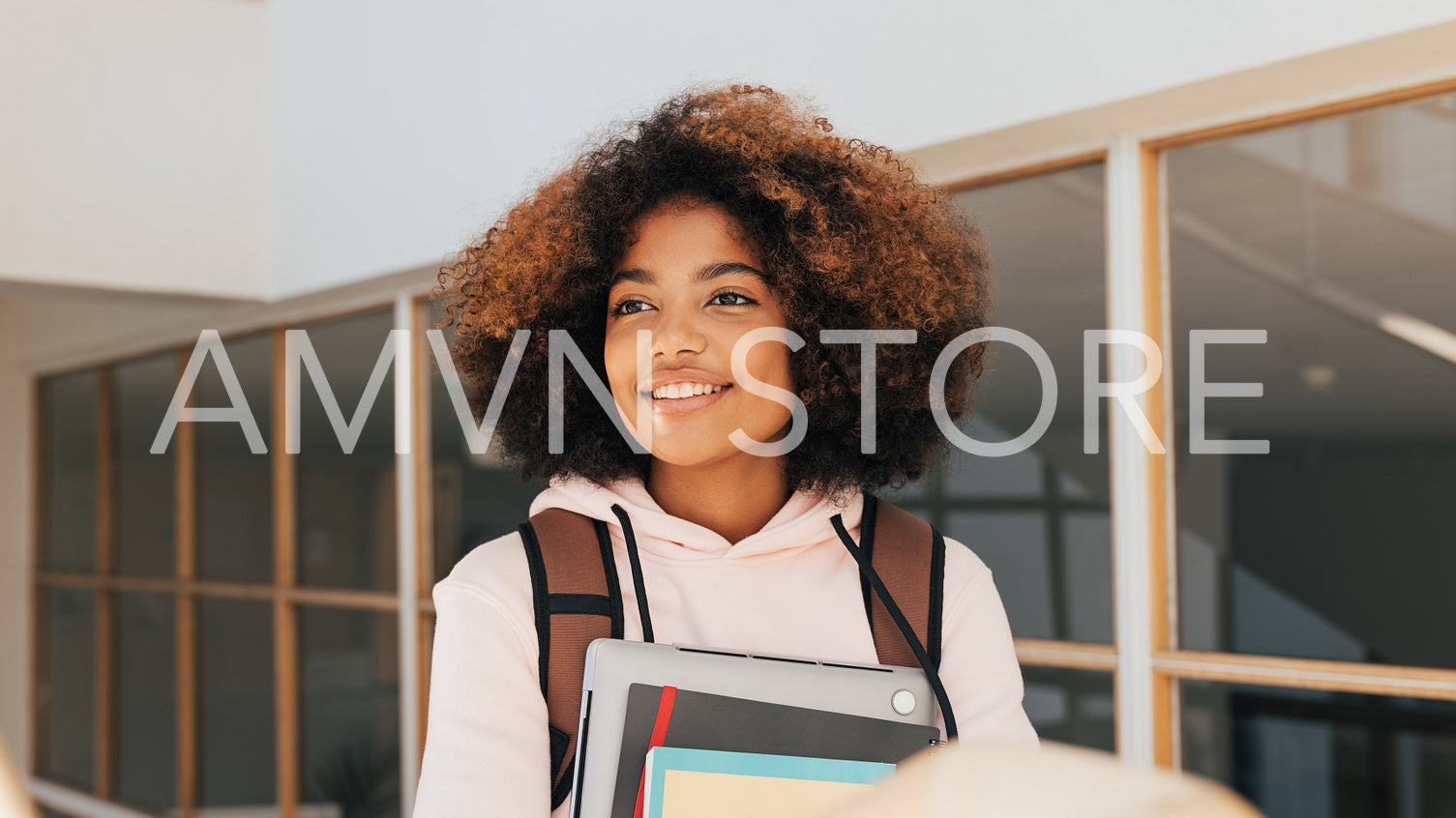 Portrait of a smiling girl with curly hair holding laptop and books while going up on staircase