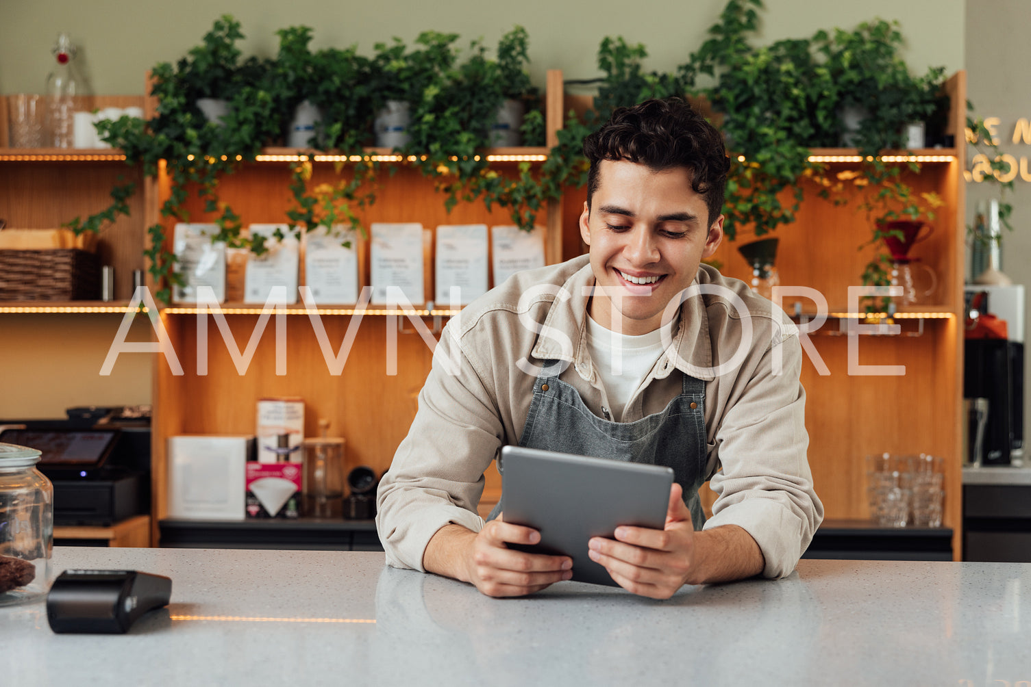 Smiling coffee shop owner at the counter with digital tablet. Male bartender in an apron leaning counter looking at a portable computer.