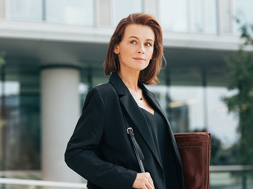 Portrait of a middle-aged businesswoman in formal clothes holding a leather folder and looking away while standing outdoors