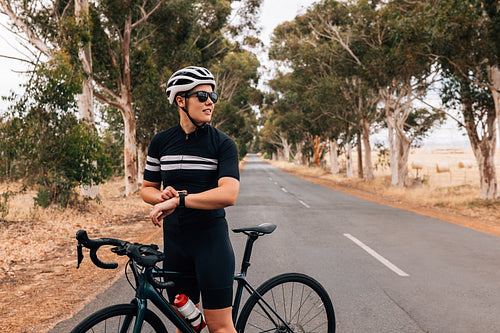 Woman cyclist with bicycle standing outdoors on countryside road and checking smartwatch