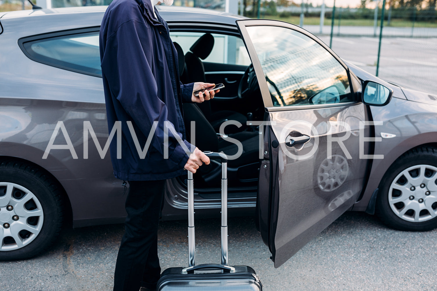 Unrecognizable man standing at car with suitcase and holding cell phone	