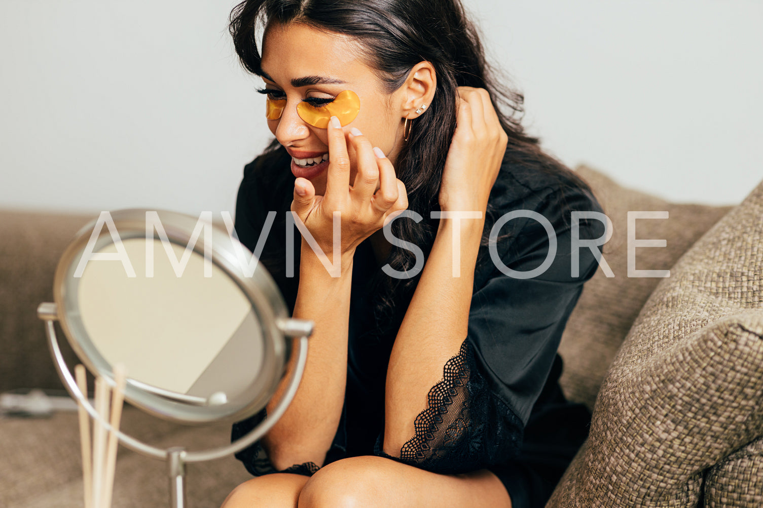 Young woman in sating robe sitting on a couch in front of a mirror applying an eye patch