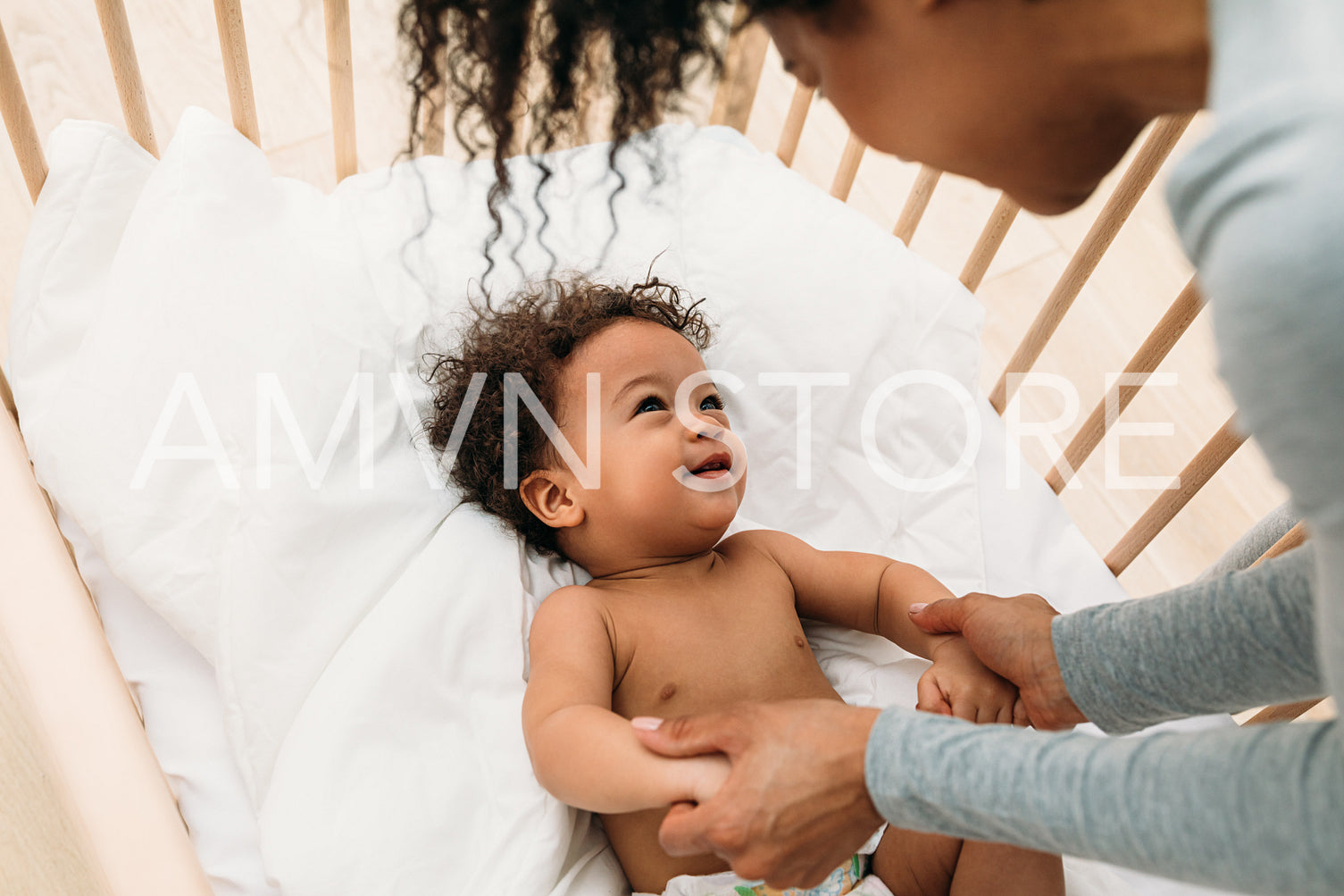 Woman leaning into a crib. Young mother looking at her son and holding hands.	