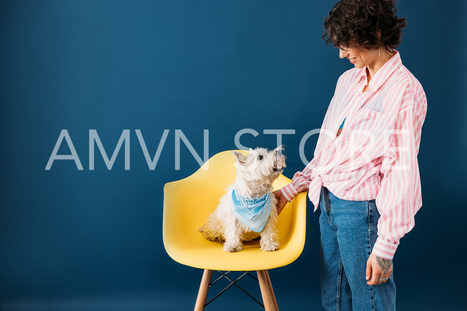 Side view of a smiling woman standing at blue backdrop with her dog who is sitting on yellow chair