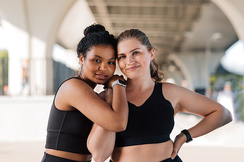 Portrait of two plus-size females in sportswear standing outdoors. Two young smiling women with similar body types hugging each other after a workout.