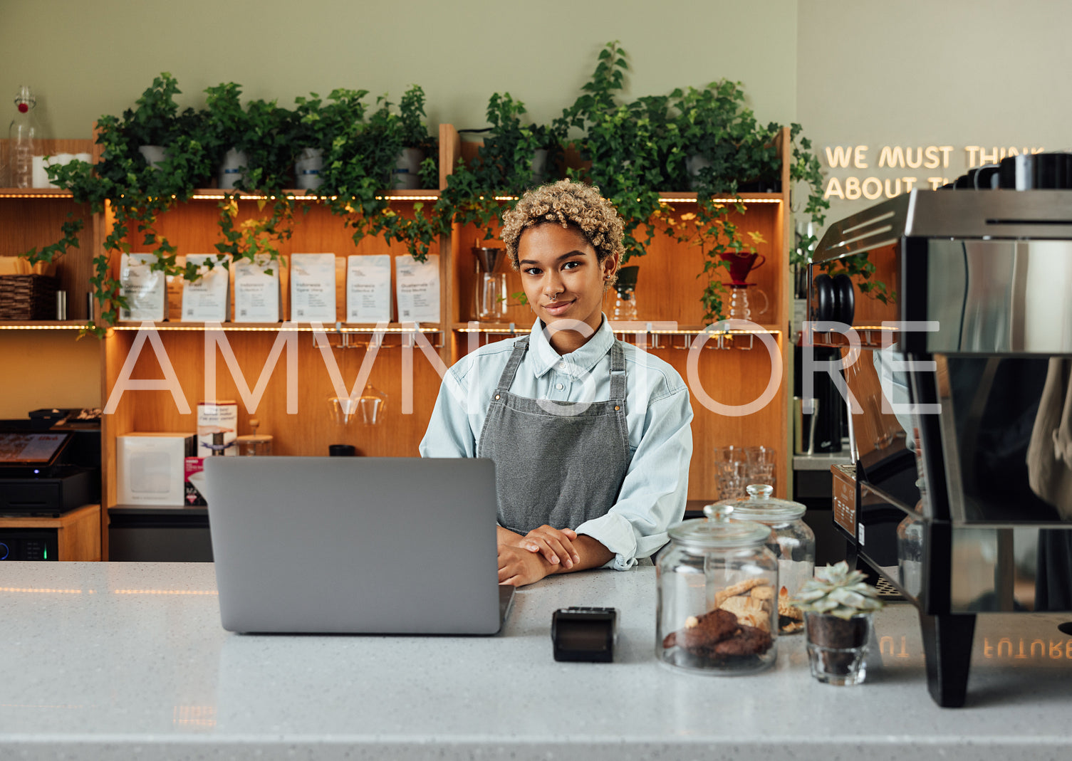 Young female entrepreneur in an apron looking at camera while standing at the counter with laptop and coffee machine