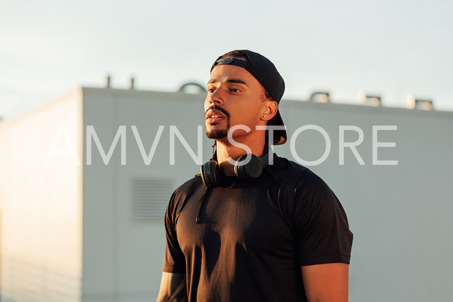 Portrait of a young confident male athlete in a cap. Athlete with wireless headphones standing on a rooftop at sunset.