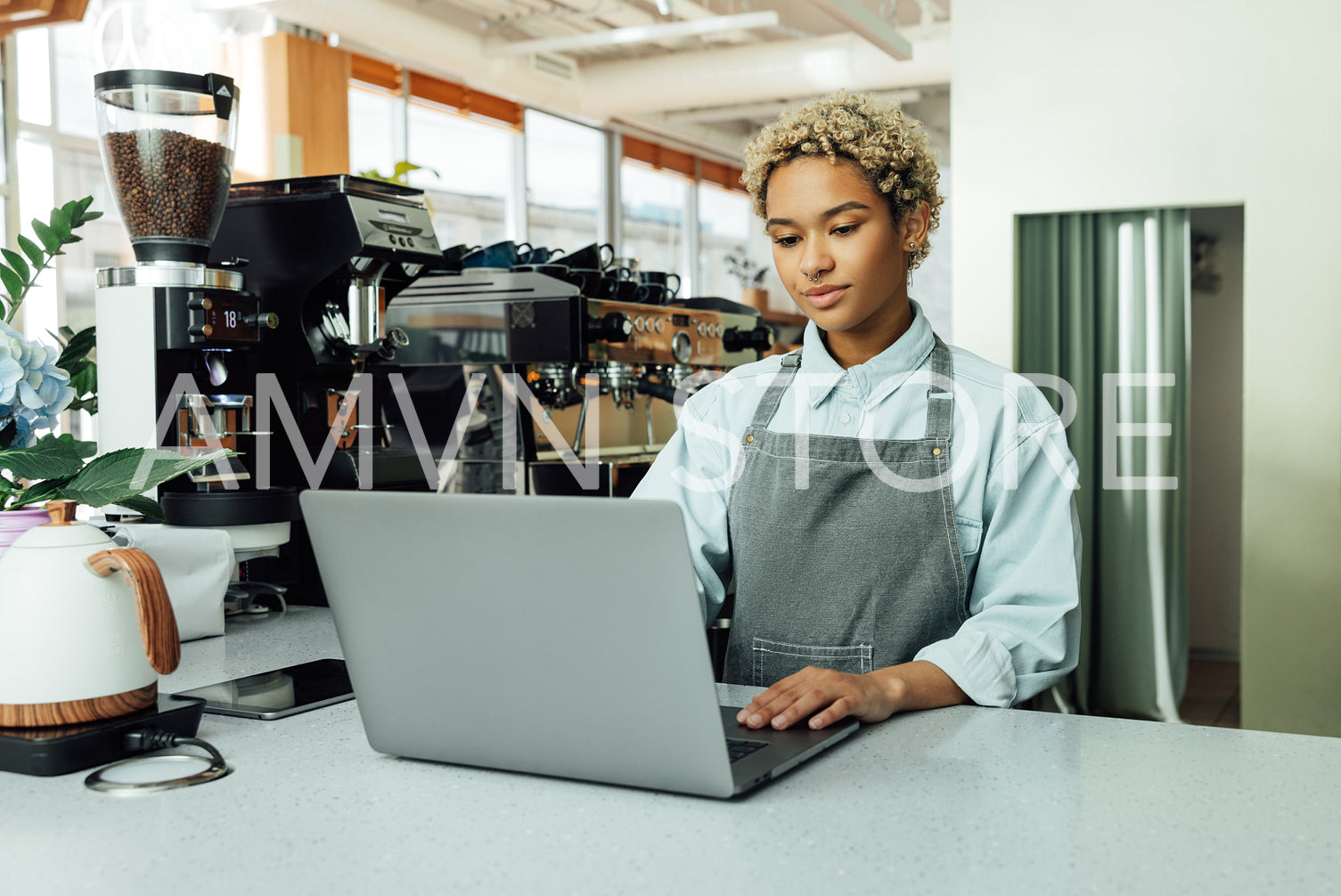 Young female barista working on laptop in a coffee shop. Woman with short hair in an apron at her small local cafe.