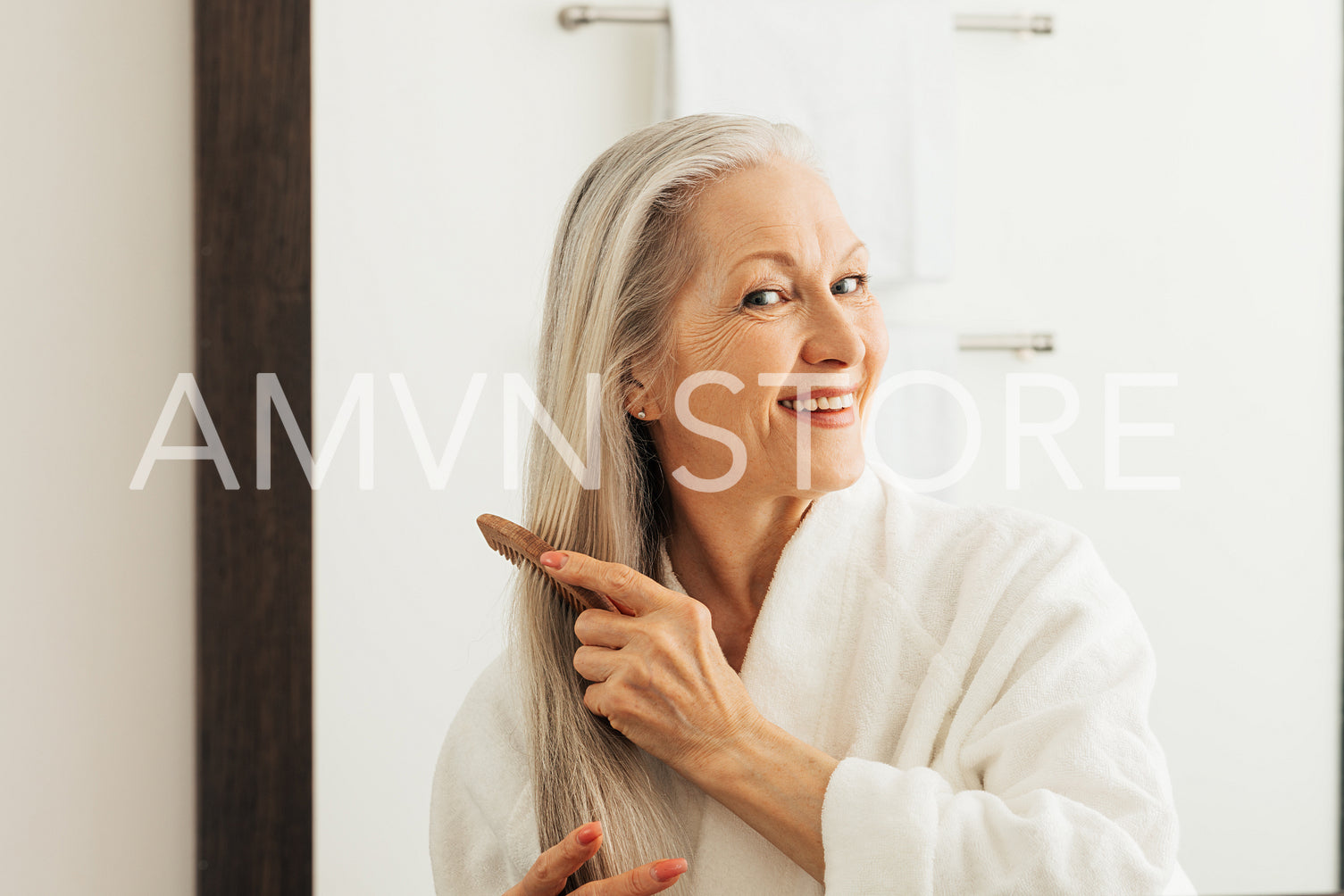 Cheerful woman combing her long hair with a wood comb in front of a bathroom mirror