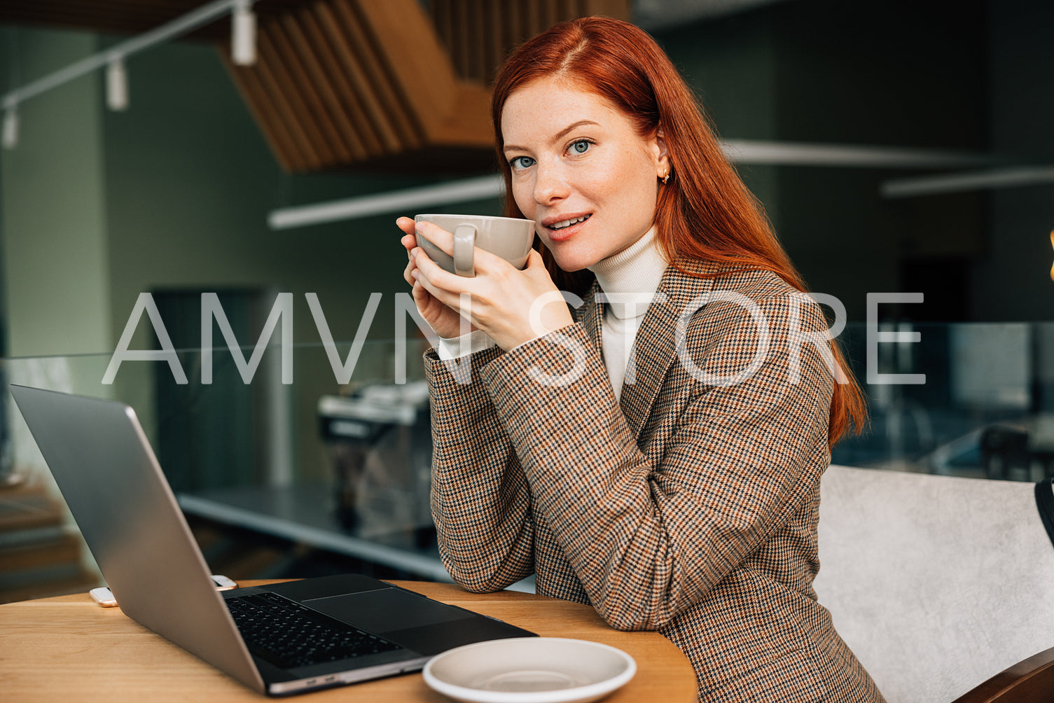 Young woman with ginger hair holding a cup. Female entrepreneur drinks coffee while working from a coffee shop.