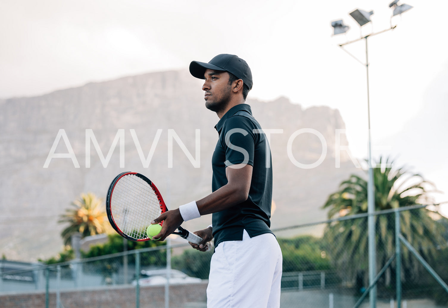 Fit man in sportswear and cap with a racket standing on tennis court. Tennis player looking away while standing on a hard court.