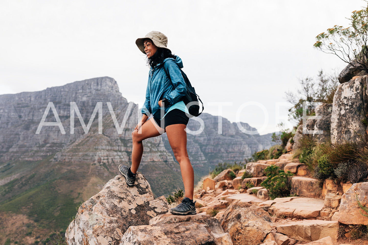 Woman wearing a hat and sports clothes relaxing during a mountain hike. Smiling female hiker looking down while standing on an edge.