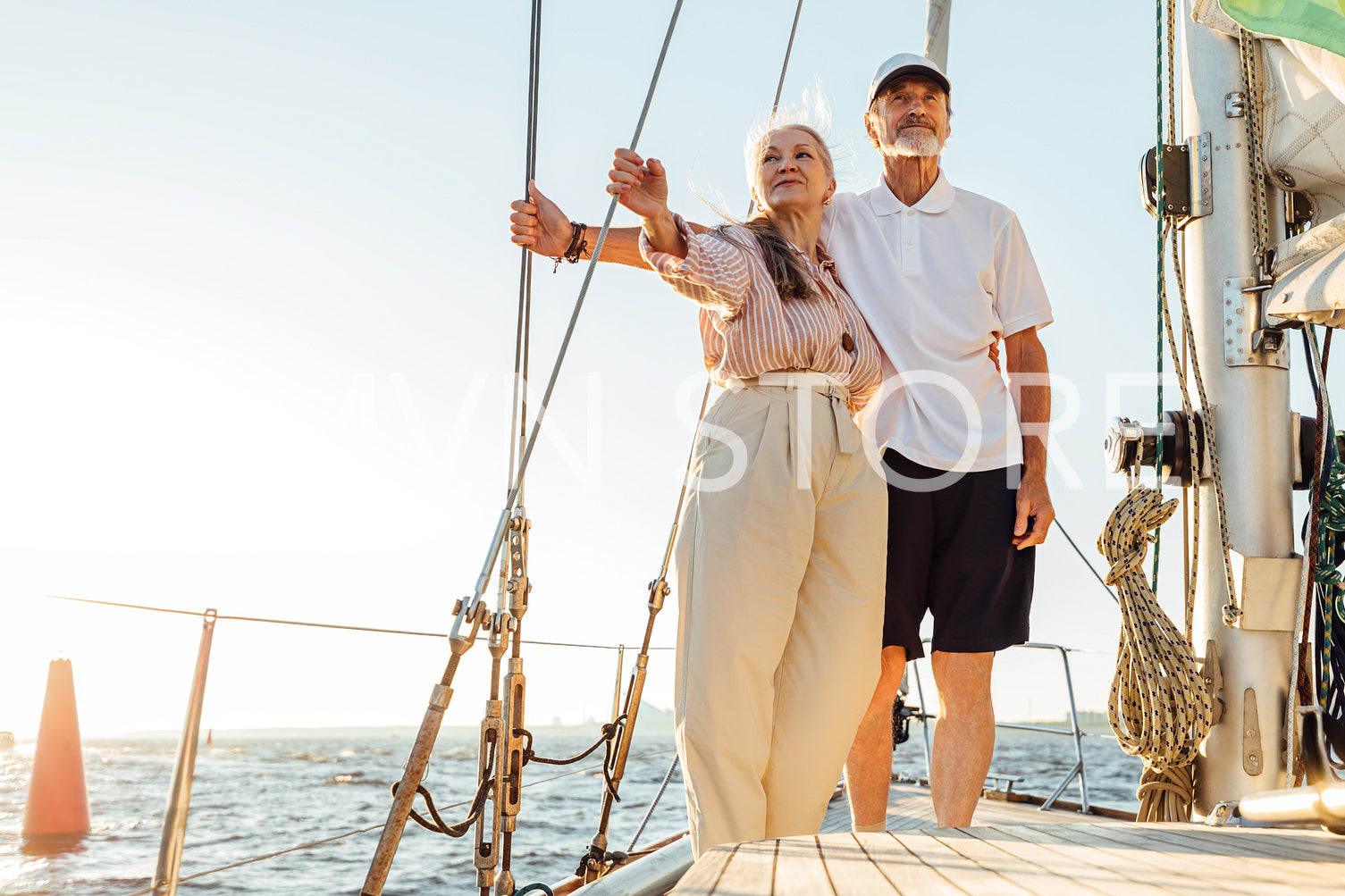 Mature husband and wife standing on a yacht and holding ropes while looking into a distance at sunset	