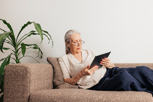 Senior woman in glasses lying on a sofa holding a digital tablet. Aged female in casuals reading from the portable computer.