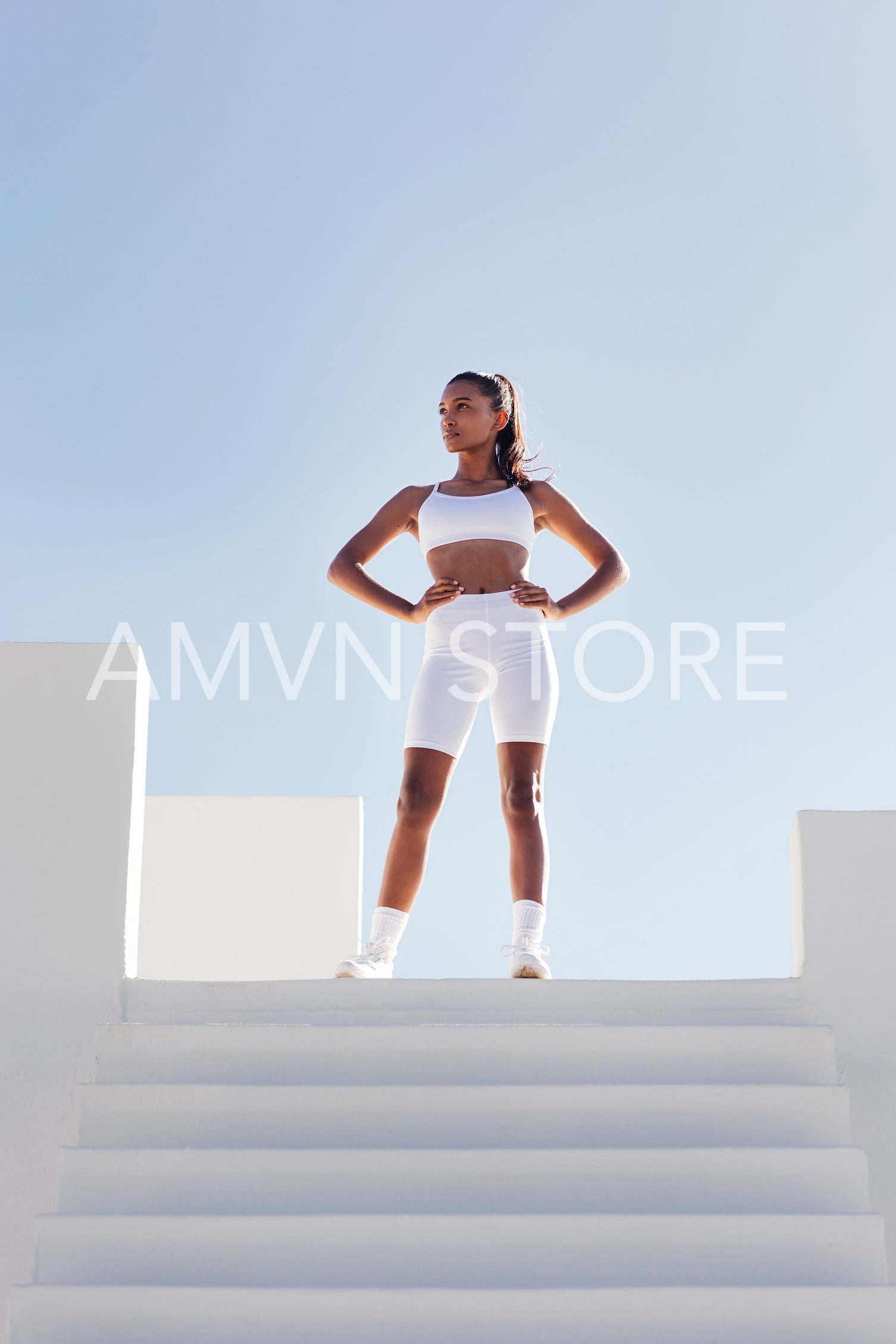 Full length of a slim confident woman looking away while standing on the top of stairs. Sportswoman in white fitness attire against skies.