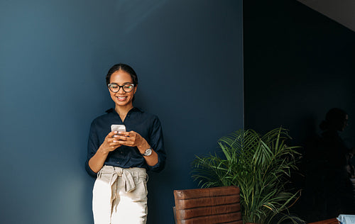 Smiling businesswoman typing on cell phone at the blue wall