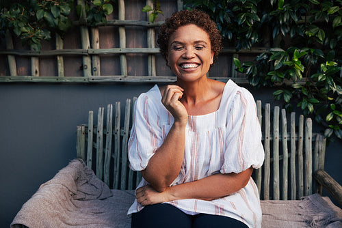 Woman laughing with closed eyes at backyard. Mature female with short hair relaxing on bench at her house.