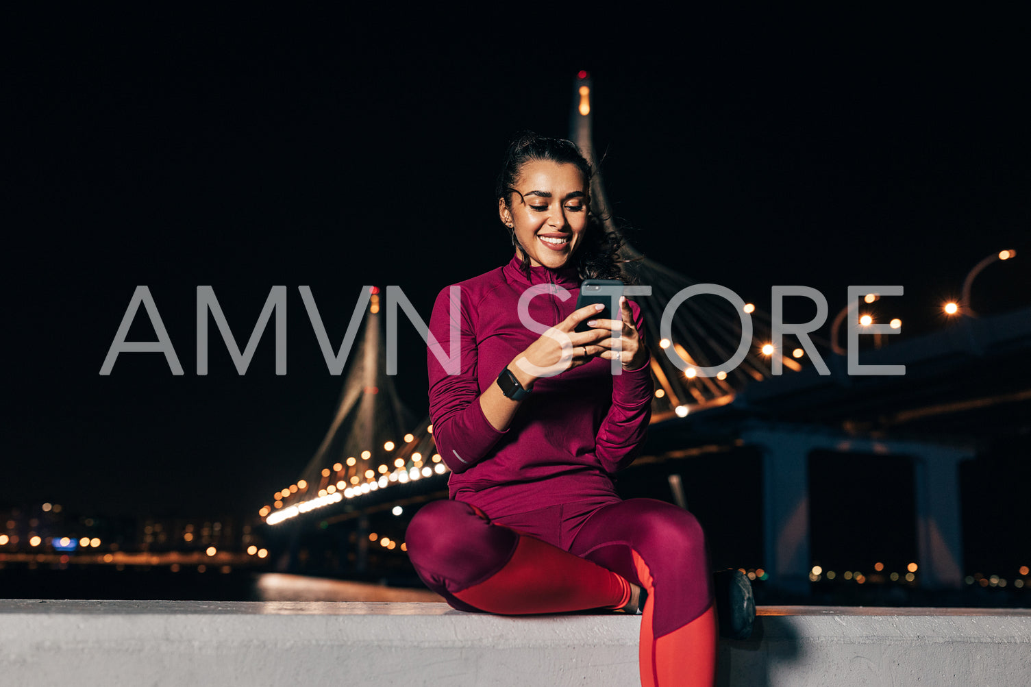 Smiling sportswoman sitting on an embankment using smartphone at night