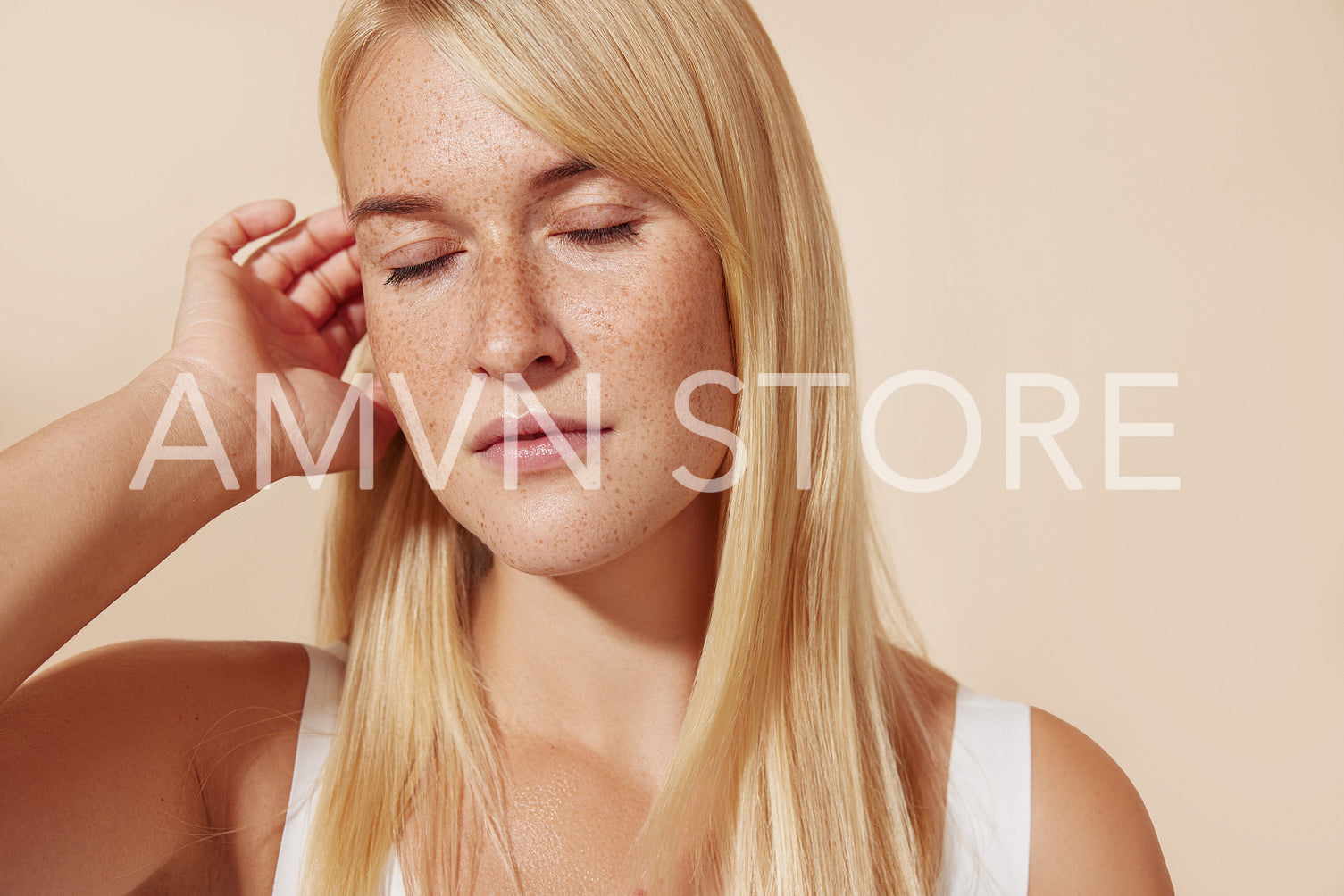 Young female with freckled skin standing with her eyes closed in studio. Blond woman adjusting hair while posing against pastel backdrop.