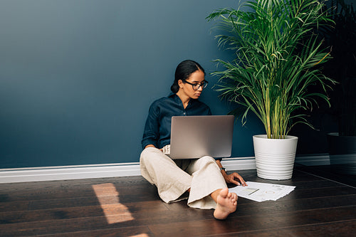 Young female entrepreneur working on laptop while sitting on floor at home. Businesswoman looking on documents while sitting at wall in living room.