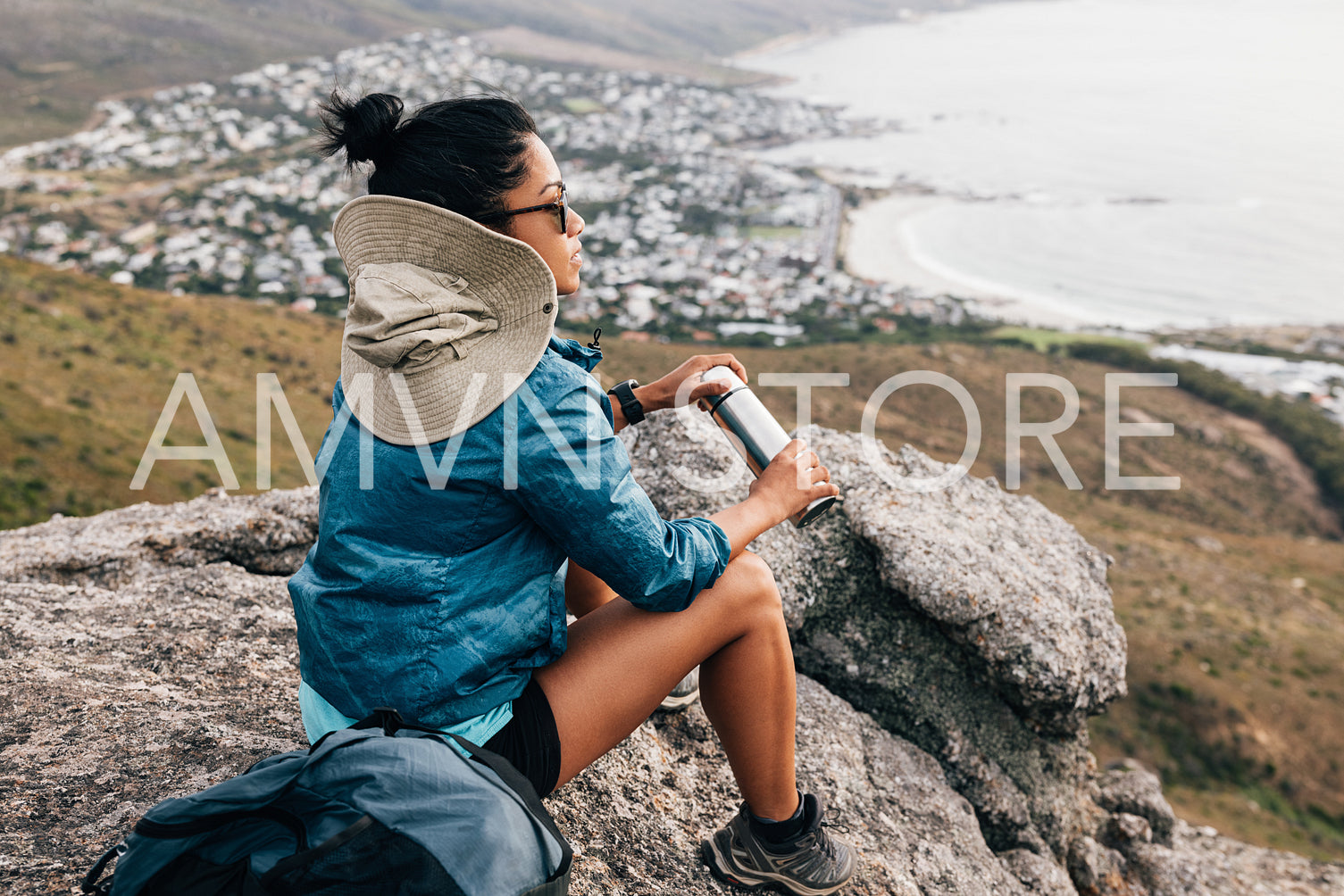 Rear view of a woman hiker in sports clothes opening a thermos while sitting on a cliff and looking at the view