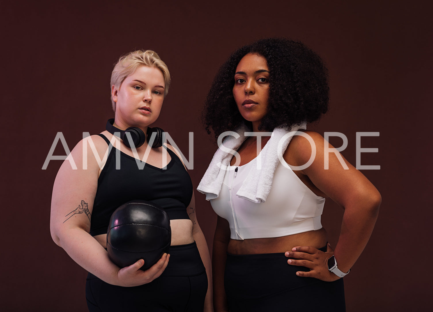 Two plus size women in sports clothes standing in studio. Young female athletes posing together against brown background.