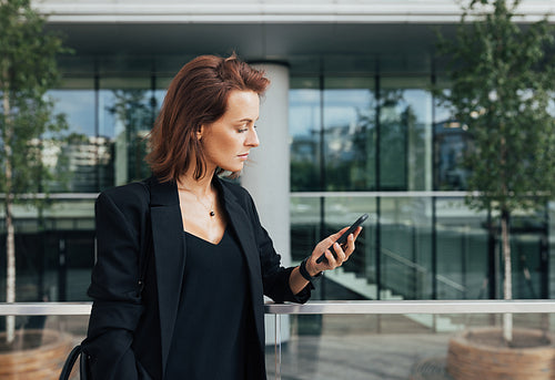 Side view of a confident businesswoman with ginger hair looking at her smartphone against an office building