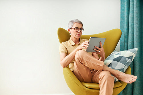 Stylish senior woman sitting at home on armchair watching content on digital tablet