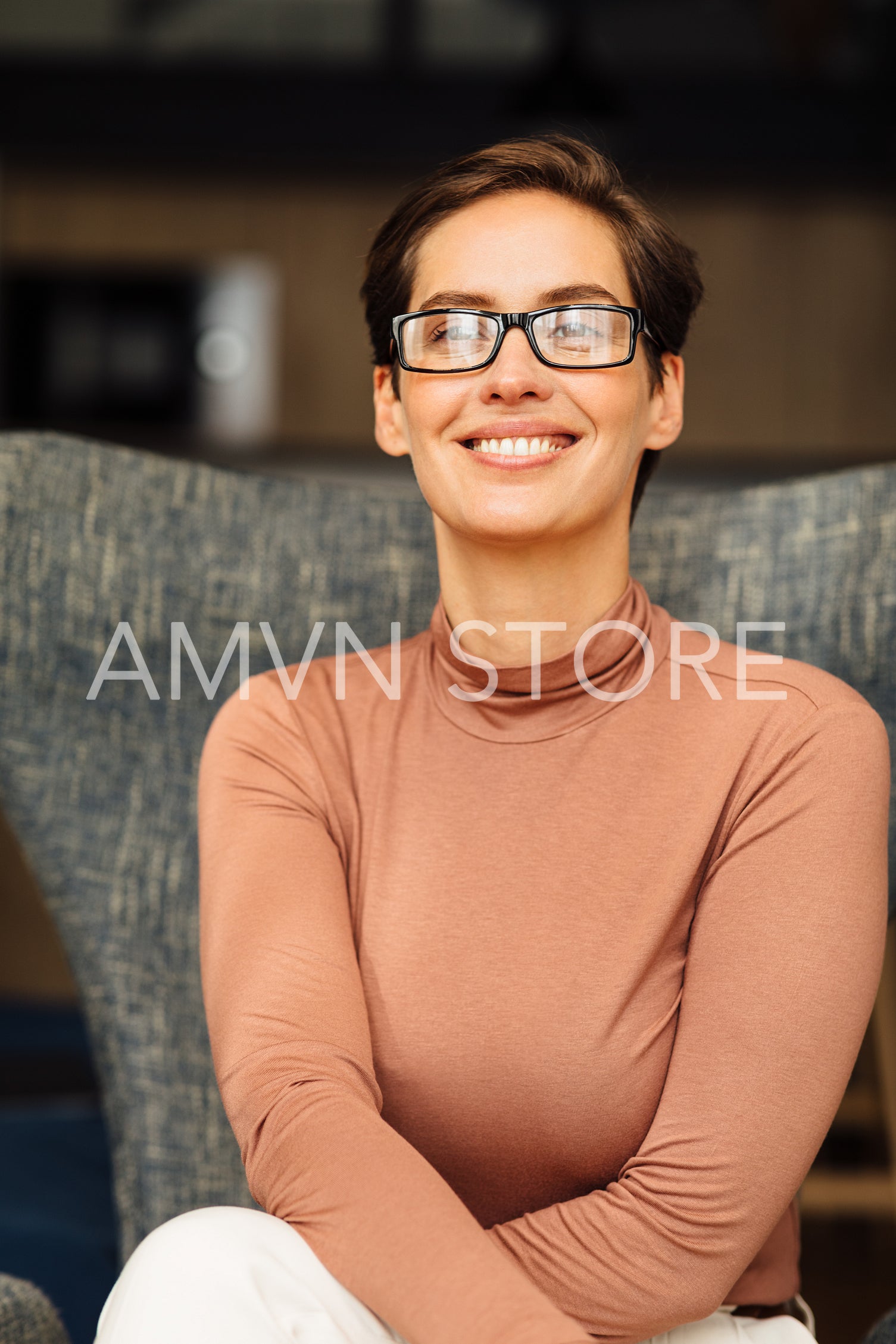 Close up portrait of a smiling woman wearing eyeglasses in an apartment. Positive female in casuals sitting on armchair.	