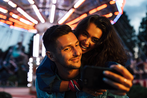 Happy female piggyback her boyfriend and takes a selfie against the carousel. Young couple having fun in an amusement park during the festival.