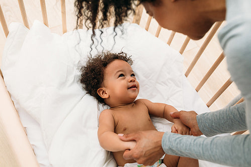 Woman leaning into a crib. Young mother looking at her son and holding hands.