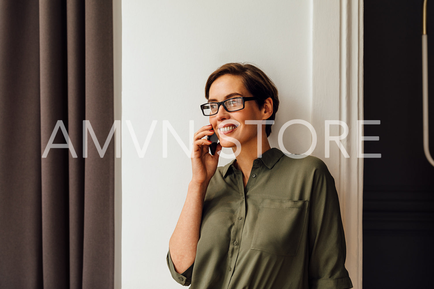 Happy businesswoman talking on mobile phone while standing at wall in rental apartment. Woman entrepreneur on business trip working from hotel room.