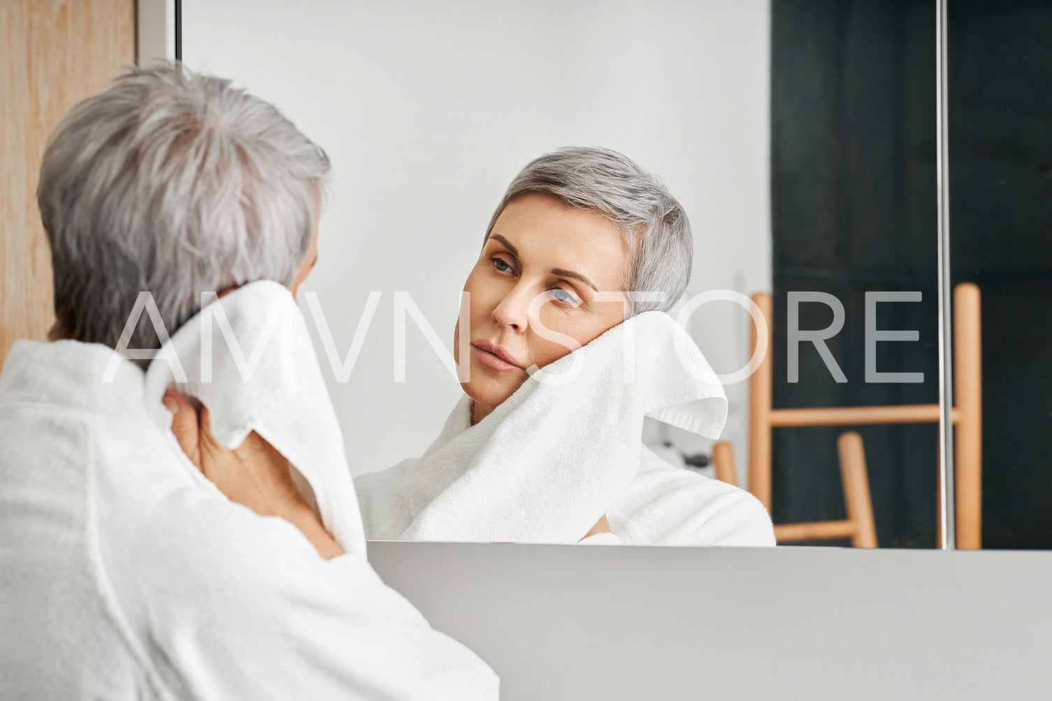 Mature woman with short grey hair looking at her reflection in bathroom mirror while wiping cheek	