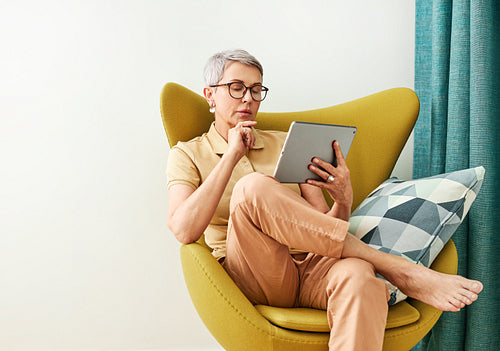 Thoughtful mature woman with closed eyes sitting at home holding a digital tablet