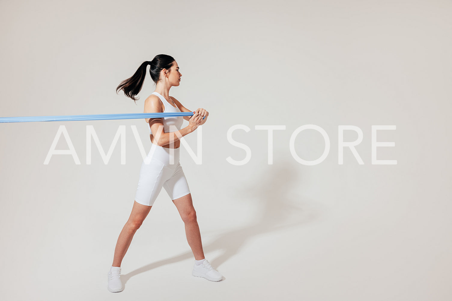 Slim female in white fitness attire pulling resistance band at the white wall in a studio