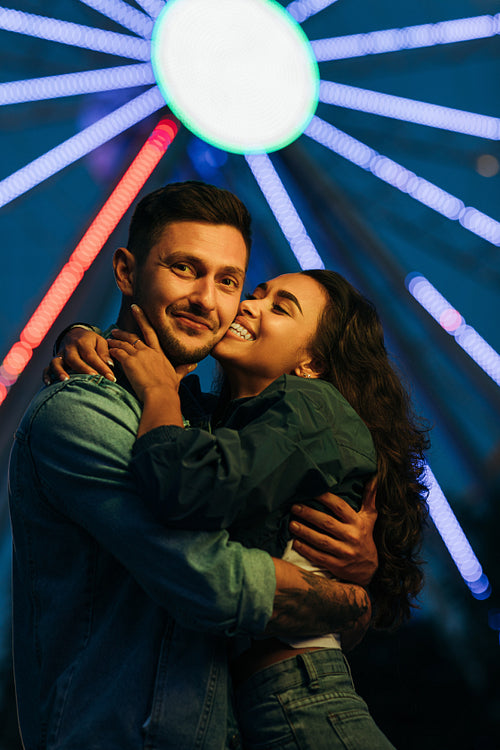 Young couple hugging and kissing against colorful lights from ferris wheel