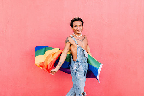 Smiling young gay man standing with pride flag. Happy guy in casuals holding LGBT flag outdoors.