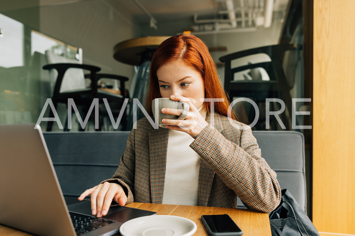 Young female with ginger hair drinking from a cup and typing on a laptop