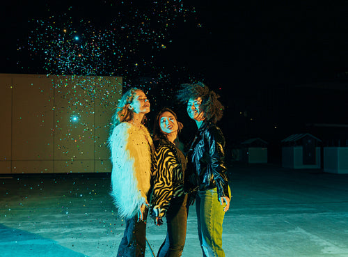 Cheerful girls standing together on a roof at night under confetti