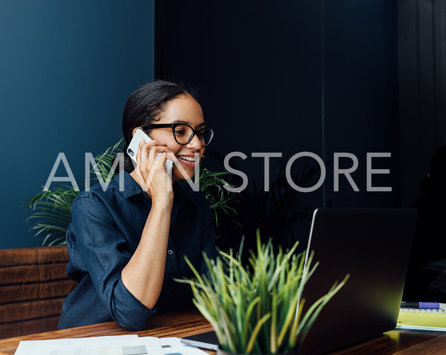 Smiling woman sitting at desk looking at laptop screen and talking over mobile phone	