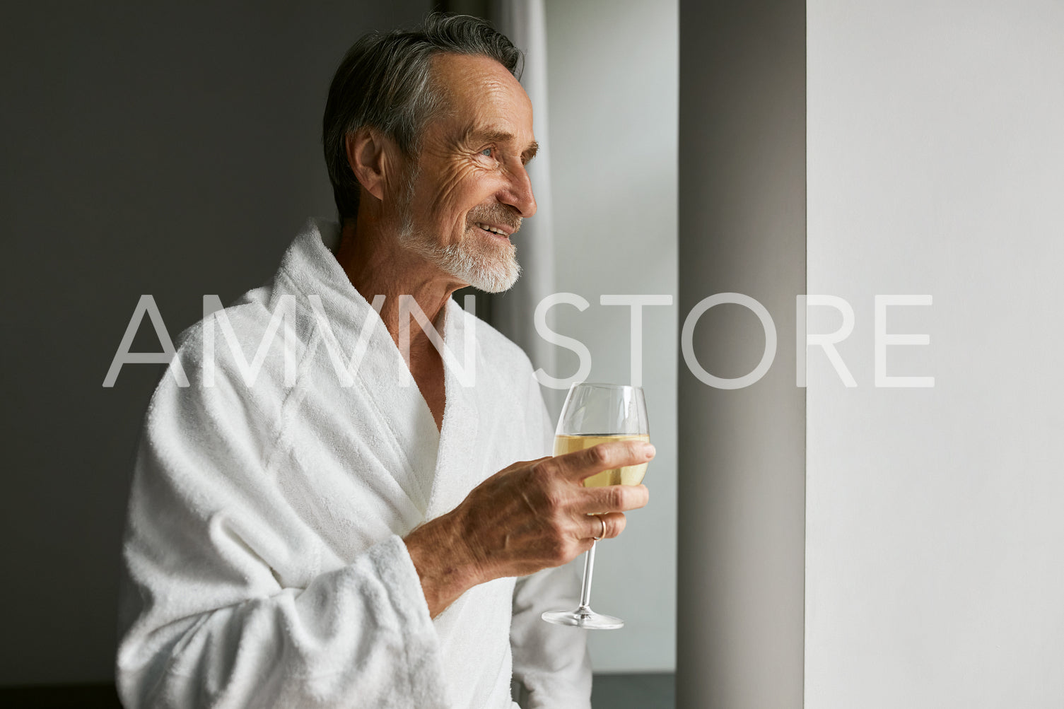 Smiling senior man with wineglass. Side view of a bearded mature male wearing a bathrobe in a hotel room looking at window.	